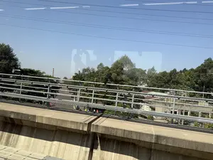 Quang cảnh nhìn từ cửa sổ của một phương tiện đang di chuyển, có thể là tàu điện trên cao. Phía trước là thành bê tông của đường ray và lan can kim loại. Bên dưới là một khu vực với nhiều cây xanh um tùm. Bầu trời trong xanh và quang đãng, có vài đường dây điện chạy ngang. Ở phía xa có thể thấy các tòa nhà và một ống khói. Có hình ảnh phản chiếu trên cửa kính.。（日本語訳）。移動中の乗り物、おそらく高架鉄道の窓から見た光景。前方には線路のコンクリート壁と金属製の手すりがある。下には、緑豊かな木々が生い茂る一帯がある。空は青く澄み渡っており、数本の電線が横切っている。ガラス窓に反射像が映っている。