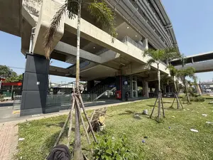 Tuyến 1. Quang cảnh bên ngoài một nhà ga tàu điện trên cao bằng bê tông vào một ngày nắng. Lối vào ga có cầu thang bộ và thang máy. Phía trước là một bãi cỏ có trồng vài cây cọ non được chống bằng cọc gỗ, trên cỏ có rác vương vãi. Một người đang đứng gần khu vực thang máy.。（日本語訳）。路線1。晴れた日の、コンクリート製の高架鉄道駅の外観。駅の入り口には階段とエレベーターがある。正面には芝生の広場があり、木の杭で支えられた若いヤシの木が数本植えられ、芝生の上にはゴミが散乱している。一人の人がエレベーターエリアの近くに立っている。