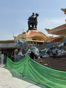 Trên tấm biển chỉ dẫn có dòng chữ: ĐIỂM SINH HOẠT VĂN HÓA, HOẠT ĐỘNG BÌNH THƯỜNG. Khung cảnh là một ngôi chùa hoặc cơ sở tín ngưỡng mang đậm kiến trúc Á Đông, có vẻ như đang trong quá trình xây dựng hoặc tu sửa. Nổi bật là một tác phẩm điêu khắc rồng lớn màu xanh lam uốn lượn dọc theo các bậc thang đi lên. Mái nhà lợp ngói đỏ, cong vút, và trên đỉnh là một chiếc lư hương bằng đồng cỡ lớn. Một vài người, có thể là công nhân, đang có mặt trên cầu thang. Phía trước, một tấm bạt màu xanh lá được dựng lên, che chắn một phần công trình.。（日本語訳）。案内板には「文化活動拠点、通常営業」という文字がある。風景は、東アジア建築の趣が色濃い寺院または信仰施設で、建設中または改修中のようである。目立っているのは、上へと続く階段に沿って曲がりくねる大きな青い龍の彫刻である。屋根は赤い瓦で葺かれ、反り上がっており、頂上には大きな銅製の香炉がある。数人の人々、おそらく作業員が、階段の上にいる。前方には、緑色のシートが立てられ、建造物の一部を覆っている。