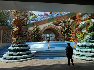 Bức ảnh chụp lối vào một công trình kiến trúc hoành tráng, mang đậm phong cách Á Đông. Nổi bật là một cầu thang rộng được sơn màu xanh lam dẫn lên một cổng vòm lớn. Hai bên cầu thang là hai cột trụ được trang trí bằng hình tượng rồng khổng lồ, nhiều màu sắc (vàng, đỏ, xanh) đang uốn lượn, với phần chân đế được tạo hình như những lớp sóng biển. Phía trên cổng là một bức tường được trang trí công phu với các bức phù điêu sặc sỡ hình cá chép, sóng nước và các họa tiết phức tạp. Có một người đàn ông mặc áo xanh, quần sáng màu, đeo ba lô đang đi lên cầu thang. Ở phía trước, một người khác trong trang phục tối màu, đội mũ và đeo ba lô, đứng trong bóng râm và nhìn về phía cầu thang. Ánh nắng mặt trời chiếu sáng rực rỡ vào công trình, tạo ra sự tương phản mạnh mẽ với khu vực bóng râm ở tiền cảnh.。（日本語訳）。写真は、東アジア風の壮大な建築物の入り口を撮影したものです。大きなアーチ門へと続く、青く塗られた広い階段が際立っています。階段の両側には、巨大で色とりどり（黄色、赤、青）の龍がとぐろを巻いている像で装飾された2本の柱があり、その土台は海の波のように形作られています。門の上には、鯉、水の波、そして複雑な模様が描かれた色鮮やかなレリーフで精巧に装飾された壁があります。青いシャツ、明るい色のズボンを履き、バックパックを背負った男性が階段を上っています。手前では、暗い色の服を着て帽子をかぶり、バックパックを背負った別の人が日陰に立ち、階段の方を見ています。太陽の光が建築物を明るく照らし、前景の日陰部分と強いコントラストを生み出しています。