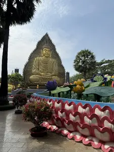 Bức ảnh chụp một pho tượng Phật lớn màu vàng đang ngồi thiền trong tư thế kiết già. Tượng Phật có vẻ mặt bình yên, thanh thản, được đặt trước một tấm phù điêu lớn hình lá bồ đề với các chi tiết chạm khắc công phu. Phía trước tượng là một đài phun nước hoặc hồ nước nhỏ, có thành được tạo hình từ những cánh sen lớn màu hồng và trắng. Bên trong hồ là những bông sen, đài sen và lá sen nhân tạo nhiều màu sắc như tím, vàng và xanh lục. Xung quanh khuôn viên có nhiều cây xanh như cây cọ, cây cảnh và những chậu hoa giấy màu hồng rực rỡ. Nền sân được lát bằng gạch đá màu xám. Ở phía xa, có một bức tượng Phật khác màu trắng. Bầu trời trong xanh và có mây.。（日本語訳）。写真には、結跏趺坐の姿勢で瞑想している大きな金色の仏像が写っている。像の前には噴水または小さな池があり、その縁はピンクと白の大きな蓮の花びらで形作られている。池の中には、紫、黄色、緑など色とりどりの造花の蓮の花、花托、葉がある。境内には、ヤシの木、観葉植物、鮮やかなピンクのブーゲンビリアの鉢植えなど、多くの緑がある。庭の地面は灰色の石のタイルで舗装されている。空は青く澄んでおり、雲がある。