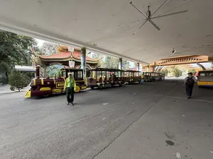 Tại một bến chờ có mái che rộng, nhiều đoàn tàu điện tham quan được xếp thành hàng dài. Các đoàn tàu có đầu kéo được thiết kế giống đầu máy xe lửa hơi nước cổ, sơn màu đỏ và vàng, kéo theo các toa xe chở khách. Một người nhân viên mặc đồng phục áo sơ mi xanh lá cây và quần tối màu đang đi bộ ở khu vực đỗ xe. Phía xa, một du khách nam đeo ba lô đang đi bộ. Phía sau là cổng vào hoặc công trình kiến trúc mang phong cách truyền thống với mái ngói đỏ. Trên trần nhà có lắp các quạt trần công nghiệp cỡ lớn.。（日本語訳）。広くて屋根のある待合所で、多くの観光用路面電車が長い列をなして並んでいます。各列車は、古い蒸気機関車のように設計された牽引車があり、赤と黄色に塗装され、客車を牽引しています。緑色のシャツと暗い色のズボンの制服を着た従業員が一人、駐車エリアを歩いています。遠くには、バックパックを背負った男性観光客が歩いています。後方には、赤い瓦屋根の伝統的な様式の門または建造物があります。天井には大型の業務用シーリングファンが設置されています。