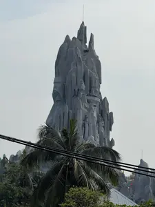 Bức ảnh chụp một công trình kiến trúc độc đáo có hình dạng một ngọn núi đá nhân tạo khổng lồ màu xám. Ngọn núi có nhiều đỉnh nhọn, lởm chởm, vươn cao lên bầu trời u ám. Trên thân núi, có thể nhận ra các hình khắc, bao gồm khuôn mặt của một người lính đội mũ và một mô hình xe tăng nhỏ, gợi liên tưởng đến một đài tưởng niệm hoặc khu di tích lịch sử. Phía trước, ngọn cây dừa và những đường dây điện đen vắt ngang qua khung hình. Bầu trời phía sau có màu xám trắng, không có nắng.。（日本語訳）。写真は、灰色の巨大な人工岩山の形をしたユニークな建築物を写している。その山は多くの尖ったごつごつした頂を持ち、曇り空に向かって高くそびえている。山肌には、帽子をかぶった兵士の顔と小さな戦車の模型を含む彫刻が見て取れ、記念碑または史跡を連想させる。手前では、ヤシの木の梢と黒い電線がフレームを横切っている。背景の空は灰白色で、日差しはない。