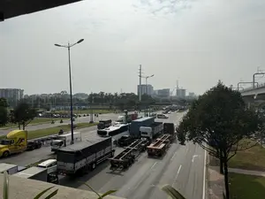 Đây là khung cảnh giao thông trên một con đường lớn, được chụp từ trên cao. Dòng xe cộ đông đúc, bao gồm rất nhiều xe tải, xe container, một chiếc xe buýt màu trắng, và một số ô tô con cùng xe máy. Bối cảnh phía sau là các tòa nhà, cây xanh, cột điện cao thế và một tuyến đường sắt trên cao. Bầu trời u ám, nhiều mây. Có thể đọc được một số chữ và số trên các phương tiện và biển báo: một tấm biển lớn màu xanh ở xa ghi 'KHU CÔNG NGHỆ CAO'. Một chiếc xe tải phủ bạt có chữ 'Phúc' và biển số '53S-049.70' viết bên hông. Một xe tải khác có biển số '53R-049.70'. Một rơ moóc màu đỏ có biển số '70R-013...'.。（日本語訳）。これは、上から撮影された大きな道路の交通風景です。車両の流れは混雑しており、多くのトラック、コンテナ車、一台の白いバス、そしていくつかの乗用車とバイクが含まれています。背景には、建物、緑の木々、高圧電線の鉄塔、そして高架鉄道があります。空はどんよりとしており、雲が多いです。車両や看板にあるいくつかの文字や数字を読み取ることができます。遠くにある大きな青い看板には「KHU CÔNG NGHỆ CAO」と書かれています。幌で覆われたトラックには「Phúc」という文字と、側面に書かれたナンバープレート「53S-049.70」があります。別のトラックにはナンバープレート「53R-049.70」があります。赤いトレーラーにはナンバープレート「70R-013...」があります。