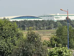 Bức ảnh chụp quang cảnh một khu công nghiệp từ xa. Ở hậu cảnh là một tòa nhà nhà máy lớn màu trắng với các chi tiết màu xanh lá cây, trên tường có logo chữ 'Nidec'. Bên cạnh là một tòa nhà khác có mái vòm cong và cửa sổ kính màu xanh lam. Ở trung cảnh là một hàng cây rậm rạp và một khu đất trống với cỏ khô. Tiền cảnh có nhiều cây cối xanh tươi. Phía bên phải có một cột đèn đường cao với thân cột màu đỏ và xanh dương. Bầu trời có màu xám nhạt.。（日本語訳）。遠くから工業団地の光景を撮影した写真。背景には、緑色のディテールがある大きな白い工場ビルがあり、壁には「Nidec」のロゴがある。隣には、湾曲したドーム屋根と青いガラス窓を持つ別の建物がある。中景には、密集した木々の列と、枯れ草のある空き地がある。前景には、青々とした木々がたくさんある。右側には、柱が赤と青の背の高い街灯がある。空は薄い灰色だ。