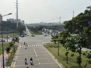 Toàn cảnh một ngã tư đường phố rộng lớn với nhiều phương tiện giao thông như xe máy, ô tô và xe tải đang di chuyển. Hai bên đường và dải phân cách được trồng nhiều cây xanh. Ở phía xa có một cây cầu vượt, các tòa nhà và những cột điện cao thế dưới bầu trời mờ sương.。（日本語訳）。広大な交差点の全景。バイク、自動車、トラックなど多くの交通車両が移動している。道路の両側と中央分離帯には多くの緑の木が植えられている。