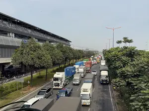 Bức ảnh chụp từ trên cao cảnh kẹt xe trên một con đường lớn với nhiều làn xe. Một hàng dài các loại xe tải, xe container và ô tô con đang nối đuôi nhau. Bên trái đường là một tòa nhà hiện đại, đó là Ga Khu Công nghệ cao, bên dưới là bãi giữ xe máy. Dọc theo con đường là những hàng cây xanh. Có thể thấy các xe tải mang nhãn hiệu THACO TRƯỜNG HẢI và các biển số xe như 50H-072.93, 86H-011.76, và 51C-952.66. Ở phía xa là cầu cạn của tuyến đường sắt trên cao. Bên lề đường phía phải, có một người đang ngồi trên ghế đẩu cạnh một chiếc xe đạp.。（日本語訳）。複数の車線がある大きな道路での交通渋滞の光景を上から撮影した写真。トラック、コンテナ車、乗用車など様々な種類の車が長い列を作って連なっている。道路の左側には近代的な建物があり、それはハイテクパーク駅で、その下はバイクの駐輪場になっている。道路に沿って緑の並木がある。道路の右側の路肩では、一人の人が自転車の隣でスツールに座っている。