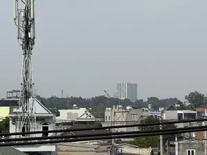 Bức ảnh chụp một khung cảnh đô thị dưới bầu trời mờ ảo. Ở phía trước, những đường dây cáp điện màu đen dày đặc chạy ngang qua. Nổi bật ở bên trái là một cột tháp viễn thông cao, có kết cấu dạng lưới. Xung quanh là các ngôi nhà dân với nhiều kiểu mái nhà khác nhau, một số nhà có bồn nước trên mái. Xen kẽ giữa các ngôi nhà là nhiều cây xanh. Ở phía xa, một tòa nhà chung cư cao tầng và một chiếc cần cẩu hiện ra lờ mờ trong không khí.。（日本語訳）。写真は、霞んだ空の下の都市の風景を写している。手前には、黒い電線が密集して横切っている。左側には、格子状の構造をした高い通信塔が目立っている。周りには様々な屋根の形の民家があり、いくつかの家は屋上に貯水タンクがある。家々の間には多くの緑の木々が点在している。