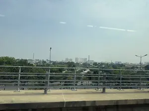 Bức ảnh chụp quang cảnh thành phố từ trên cao, có lẽ là từ một chuyến tàu điện trên cao hoặc một cây cầu. Phía trước là lan can an toàn bằng kim loại. Nhìn qua lan can là một khu vực rộng lớn với nhiều cây xanh rậm rạp. Xa hơn nữa là các tòa nhà dân cư và các tòa nhà chung cư cao tầng. Có thể thấy các cột điện cao thế và đường dây điện chằng chịt trên nền trời. Bầu trời trong xanh nhưng có vẻ hơi mờ ảo do sương mù hoặc nắng gắt.。（日本語訳）。高所から撮影された街の風景写真で、おそらく高架鉄道か橋の上から撮られたものでしょう。手前には金属製の安全柵があります。柵の向こうには、緑豊かな木々が生い茂る広大なエリアが見えます。そのさらに向こうには、住宅や高層マンションが建ち並んでいます。空を背景に、高圧電線の鉄塔や電線が複雑に交差しているのが見えます。空は青く澄んでいますが、霧または強い日差しのせいか、少し霞んで見えます。