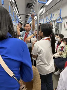 Bức ảnh chụp cảnh bên trong một toa tàu điện đông đúc. Ở trung tâm là một người đàn ông trung niên mặc áo sơ mi xanh nhạt, quàng khăn đỏ, đang đứng vịn tay nắm và nói chuyện với một nhóm học sinh. Các em học sinh, nhiều em cũng quàng khăn quàng đỏ, đang đứng và ngồi xung quanh, lắng nghe. Một số học sinh mặc đồng phục. Khung cảnh cho thấy đây có thể là một chuyến đi dã ngoại của thầy và trò trên phương tiện công cộng.。（日本語訳）。写真は混雑した電車の一車両の内部を写しています。中央には、水色のシャツを着て赤いスカーフを巻き、吊り革につかまって生徒の一団と話している中年の男性がいます。生徒たちは、多くが同じく赤いスカーフを巻いており、周りに立ったり座ったりして聞いています。一部の生徒は制服を着ています。この光景は、先生と生徒たちが公共交通機関を利用して遠足に出かけている様子を示しているのかもしれません。