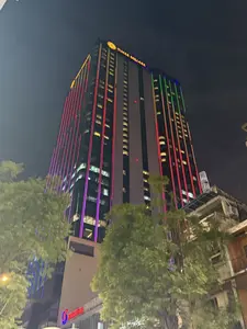 Bức ảnh chụp một tòa nhà cao tầng vào ban đêm, nổi bật với hệ thống đèn LED nhiều màu sắc chạy dọc theo mặt tiền, bao gồm các màu đỏ, tím, xanh lá và vàng. Trên đỉnh tòa nhà có dòng chữ 'TIMES SQUARE' màu vàng. Phía trước tòa nhà có những hàng cây xanh. Một số cửa sổ của tòa nhà vẫn sáng đèn dưới bầu trời đêm. Ở phía dưới có thể thấy logo của Benthanh Group.。（日本語訳）。夜の高層ビルの写真で、ファサードに沿って走る赤、紫、緑、黄色を含む多色のLEDライトシステムが際立っています。ビルの頂上には黄色の「TIMES SQUARE」という文字があります。ビルの前には緑の並木があります。夜空の下、ビルのいくつかの窓はまだ明かりが灯っています。下部にはBenthanh Groupのロゴが見えます。