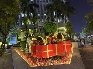 Khung cảnh ban đêm tại một thành phố, nổi bật với cụm trang trí gồm nhiều hộp quà lớn màu đỏ thắt nơ vàng và được chiếu sáng rực rỡ. Xung quanh các hộp quà là bồn cây với những cây lá đỏ và dải đèn vàng ấm áp. Phía sau là tiểu cảnh hòn non bộ, cây xanh và cây cọ. Ở hậu cảnh là một tòa nhà cao tầng hiện đại. Bên phải có một vài người đang đi dạo trên vỉa hè, tạo nên một không khí lễ hội.。（日本語訳）。ある都市の夜景。金色のリボンが結ばれ、きらびやかにライトアップされた大きな赤いギフトボックスのオブジェが際立っている。ギフトボックスの周りには、赤い葉の植物と暖かい黄色のイルミネーションが飾られた植え込みがある。その後ろには、築山、緑の木々、ヤシの木のミニチュア風景がある。背景には近代的な高層ビルがある。右側では、数人が歩道を散策しており、祝祭的な雰囲気を醸し出している。