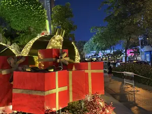 Khung cảnh một con đường vào ban đêm, được trang trí rực rỡ cho mùa lễ hội. Nổi bật ở phía trước là các hộp quà Giáng sinh mô hình cỡ lớn màu đỏ, được thắt nơ màu be và vàng. Phía sau chúng là những quả châu trang trí khổng lồ được viền bằng dây đèn LED phát sáng màu vàng ấm. Xung quanh là cây cối xanh tươi. Ở phía xa, có một người đang đứng trên vỉa hè, cùng với cây cối, xe cộ và ánh đèn từ các tòa nhà, tạo nên một không khí nhộn nhịp của thành phố về đêm.。（日本語訳）。祝祭シーズンに向けて華やかに飾られた、夜の道の風景。前景で目立っているのは、ベージュと金色のリボンが結ばれた、大きな赤い模型のクリスマスプレゼントの箱である。その後ろには、暖かい黄色の光を放つLEDライトストリングで縁取られた、巨大な装飾用のオーナメントボールがある。周りには青々とした木々がある。遠くには、歩道に一人の人が立っており、木々、車、建物からの光と共に、夜の街の賑やかな雰囲気を醸し出している。