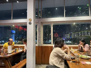 Một nhóm người đang ăn tối trong một nhà hàng vào ban đêm. Qua các ô cửa sổ lớn, có thể thấy quang cảnh thành phố với nhiều tòa nhà cao tầng sáng đèn. Bên trong, không gian được bài trí bằng bàn ghế gỗ. Có hai nhóm khách: một bàn gồm hai người đàn ông đang trò chuyện, trong đó một người mặc áo polo màu vàng; bàn còn lại là một nhóm phụ nữ đang ăn uống vui vẻ. Trên bàn ăn có nhiều món nướng, salad và đồ uống. Trên bàn của hai người đàn ông có một hộp giấy ghi chữ "10 điểm".。（日本語訳）。あるグループが夜、レストランで夕食をとっている。店内は、木製のテーブルと椅子で設えられている。客は2つのグループがいる。1つのテーブルでは男性2人が話しており、そのうち1人は黄色のポロシャツを着ている。もう1つのテーブルでは女性のグループが楽しそうに食事をしている。食事のテーブルには、多くの焼き物、サラダ、飲み物がある。男性の2人組のテーブルの上には、「10 điểm」と書かれた紙箱がある。