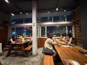 Bên trong một nhà hàng vào buổi tối, có hai nhóm khách đang ngồi ăn uống tại hai chiếc bàn gỗ dài. Không gian quán có sàn bê tông và tường ốp gỗ, nhìn ra khung cảnh thành phố về đêm qua những ô cửa sổ lớn. Một nhóm gồm hai người đàn ông và một phụ nữ đang ngồi ở bàn bên trái, trong khi bàn bên phải là một nhóm gồm ba người phụ nữ đang trò chuyện và dùng bữa. Trên bàn có nhiều món ăn, đồ uống và thực đơn. Không khí trong quán trông ấm cúng và mọi người đang thư giãn, tận hưởng bữa tối.。（日本語訳）。夜のとあるレストランの中、2つの長い木製のテーブルで2組の客が飲食をしています。店内はコンクリートの床と木製の壁で、大きな窓からは夜の街の景色が見えます。左側のテーブルには男性2人と女性1人のグループが座っており、一方、右側のテーブルでは女性3人のグループが会話をしながら食事をしています。テーブルの上にはたくさんの料理、飲み物、そしてメニューがあります。店内の雰囲気は居心地が良さそうで、皆リラックスしてディナーを楽しんでいます。