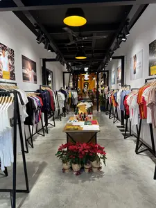 Không gian bên trong một cửa hàng thời trang với thiết kế hiện đại. Cửa hàng có dạng hình ống, dài và hẹp. Hai bên lối đi là các dãy giá treo quần áo, trưng bày nhiều mẫu áo thun và áo nỉ với màu sắc đa dạng. Trần nhà màu đen, được lắp đặt hệ thống đèn rọi và các đèn treo lớn màu vàng. Sàn nhà được làm bằng bê tông mài. Trên các bức tường trắng có treo những tấm áp phích lớn hình người mẫu. Ở giữa cửa hàng có một chiếc bàn gỗ trưng bày các sản phẩm gấp gọn và vật trang trí, bao gồm một bức tượng nhỏ màu vàng đội nón lá. Phía trước lối vào có đặt một vài chậu hoa trạng nguyên màu đỏ rực rỡ. Ở phía xa trong cửa hàng có vài khách hàng đang lựa chọn sản phẩm.。（日本語訳）。モダンなデザインのファッション店の内部空間。店は筒状で、細長い形をしている。通路の両側には衣類ラックが並び、様々な色のTシャツやスウェットシャツが多数陳列されている。床は研磨コンクリートでできている。白い壁にはモデルの大きなポスターが飾られている。店の中央には、折りたたまれた商品や装飾品を陳列する木製のテーブルがあり、ノンラーをかぶった小さな黄色い像も含まれている。店の奥の方では、数人の客が商品を選んでいる。