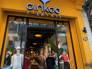 Mặt tiền của một cửa hàng quần áo tên là 'Ginkgo T-SHIRTS' được sơn màu vàng rực rỡ. Số nhà 107 được gắn trên tường bên phải. Qua ô cửa kính lớn, có thể thấy bên trong cửa hàng trưng bày nhiều ma-nơ-canh mặc các loại áo phông và áo ba lỗ. Một ma-nơ-canh nam mặc áo ba lỗ trắng đội mũ lưỡi trai, bên cạnh là một ma-nơ-canh nữ mặc áo không tay màu đỏ có họa tiết hoa lá và đội mũ tai bèo. Sâu bên trong là không gian cửa hàng với nhiều dãy quần áo được treo trên giá, dưới ánh đèn vàng ấm áp, tạo cảm giác hiện đại và thân thiện.。（日本語訳）。「Ginkgo T-SHIRTS」という名前の衣料品店のファサードは、鮮やかな黄色に塗られている。107番地が右側の壁に取り付けられている。白いタンクトップを着てキャップをかぶった男性マネキン、その隣には花柄の赤いノースリーブを着てバケットハットをかぶった女性マネキンがいる。奥には、暖かい黄色の照明の下、ラックに掛けられた多くの衣類の列がある店の空間が広がっており、モダンで親しみやすい感覚を生み出している。