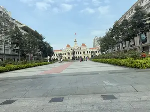 Bức ảnh chụp một quảng trường đi bộ rộng lớn, nhìn thẳng về phía tòa nhà Ủy ban Nhân dân Thành phố Hồ Chí Minh. Tòa nhà mang kiến trúc cổ điển của Pháp với màu vàng đặc trưng, ở giữa có một tháp đồng hồ và trên đỉnh là lá cờ đỏ sao vàng của Việt Nam đang tung bay. Phía trước tòa nhà là tượng đài Chủ tịch Hồ Chí Minh. Quảng trường được lát đá sạch sẽ, hai bên là hàng cây xanh và các bồn hoa được chăm sóc cẩn thận. Có một vài người dân và du khách đang đi dạo, tham quan và chụp ảnh. Bầu trời trong xanh, quang đãng, tạo nên một khung cảnh đẹp và bình yên tại trung tâm thành phố.。（日本語訳）。写真は、ホーチミン市人民委員会庁舎をまっすぐ望む、広大な歩行者広場を写している。庁舎は特徴的な黄色をしたフランスの古典建築様式で、中央には時計塔があり、頂上ではベトナムの金星紅旗がはためいている。庁舎の前にはホー・チ・ミン主席の像がある。広場はきれいに石畳が敷かれ、両側には緑の並木と手入れの行き届いた花壇がある。何人かの市民や観光客が散歩したり、見学したり、写真を撮ったりしている。空は青く澄み渡り、都心に美しく平和な光景を作り出している。