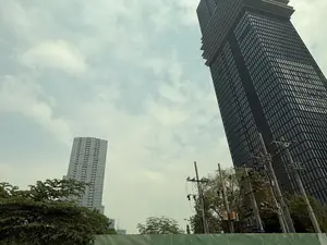 Bức ảnh chụp từ góc thấp hướng lên hai tòa nhà cao tầng dưới bầu trời nhiều mây. Nổi bật ở bên phải là một tòa nhà chọc trời rất cao, màu sẫm với mặt tiền bằng kính, phần đỉnh có kiến trúc giật cấp. Ở phía xa bên trái là một tòa nhà cao tầng khác có màu sáng hơn. Phía dưới là những ngọn cây xanh, các cột điện với dây điện chằng chịt và một hàng rào tôn màu xanh.。（日本語訳）。曇り空の下、2つの高層ビルをローアングルから見上げて撮影した写真。右側に目立つのは、非常に高い暗色の超高層ビルで、ガラス張りのファサードを持ち、頂上部分は段状の建築になっている。左の遠方には、より明るい色の別の高層ビルがある。下の方には、緑の木々の梢、複雑に絡み合った電線を持つ電柱、そして青いトタンのフェンスがある。