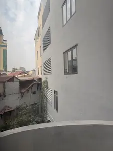 Bức ảnh chụp quang cảnh các tòa nhà san sát nhau trong một khu đô thị, nhìn từ trên cao. Bên phải là một tòa nhà hiện đại, nhiều tầng với bức tường màu xám, có các cửa sổ kính và ô thông gió. Bên trái là những ngôi nhà cũ hơn với mái ngói đỏ và mái tôn. Giữa các tòa nhà có một bụi tre đang vươn lên. Bầu trời có nhiều mây. Phía trước là một phần của lan can bê tông.。（日本語訳）。高い場所から見た、ある都市部で建物が密集している様子の写真。右側には、灰色の壁、ガラス窓、換気口がある近代的な高層ビルがある。左側には、赤い瓦屋根やトタン屋根の古い家々がある。建物の間には、上に伸びている竹の茂みがある。空は雲が多い。手前には、コンクリート製の手すりの一部がある。