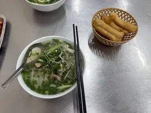 Trên một chiếc bàn ăn bằng inox, có một tô phở gà nóng hổi. Trong tô phở trắng, có thể thấy rõ bánh phở, những miếng thịt gà xé, và rất nhiều hành lá được thái nhỏ rắc lên trên. Một chiếc thìa inox nằm trong tô và một đôi đũa màu đen được gác trên miệng tô. Bên cạnh tô phở là một chiếc rổ nhỏ bằng nhựa màu be, bên trong đựng những chiếc quẩy chiên vàng giòn. Ở phía trên của bức ảnh, có một phần của một tô phở khác và một khay đựng ớt thái lát.。（日本語訳）。ステンレス製の食卓の上に、熱々の鶏肉のフォーが一杯あります。白いフォーの丼の中には、フォーの麺、細かく裂いた鶏肉、そしてその上に振りかけられたたくさんの刻みネギがはっきりと見えます。丼の中にはステンレス製のスプーンが一本あり、丼の縁には黒い箸が一膳置かれています。フォーの丼の隣には、ベージュ色の小さなプラスチック製の籠があり、中には黄金色に揚げられたサクサクのクワイ（揚げパン）が入っています。写真の上部には、別のフォーの丼の一部と、スライスされた唐辛子が入ったトレイがあります。