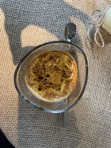 Một ly cà phê được chụp từ góc nhìn trên xuống. Ly được làm bằng thủy tinh trong suốt, có hình dáng hơi méo. Trên bề mặt cà phê là một lớp bọt kem dày màu vàng, được rắc bột màu nâu sẫm, có thể là bột ca cao hoặc quế. Ly cà phê được đặt trên một tấm lót bàn dệt từ sợi thô màu be. Bên cạnh ly có một chiếc thìa kim loại và ở góc trên bên phải là một phần của cây nến được quấn bằng dây thừng.。（日本語訳）。一杯のコーヒーが、真上からの視点で撮影されている。グラスは透明なガラスでできており、形は少し歪んでいる。コーヒーの表面には、黄色い厚いクリームの泡の層があり、その上にはココアパウダーかシナモンと思われる濃い茶色の粉が振りかけられている。