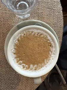 Một tách cà phê được chụp từ trên xuống. Tách cà phê màu trắng đặt trên một chiếc đĩa màu xanh nhạt. Bề mặt cà phê có một lớp bọt dày, màu nâu ở giữa và viền bọt trắng xung quanh. Tách cà phê được đặt trên một tấm lót bằng vải bố màu nâu. Ở góc trên bên trái có một phần của ly nước thủy tinh.。（日本語訳）。一杯のコーヒーが真上から撮影されている。コーヒーの表面には厚い泡の層があり、中央は茶色で、周りは白い泡で縁取られている。左上隅には、ガラスのコップの一部がある。