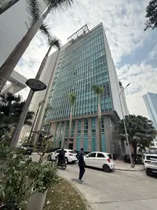 Bức ảnh chụp một tòa nhà văn phòng cao tầng, hiện đại với mặt tiền bằng kính, được chụp từ góc thấp hướng lên trời. Bầu trời có mây trắng. Xung quanh tòa nhà có những cây cọ cao. Dưới đường có một vài chiếc ô tô đang đỗ và một người đàn ông đang đi bộ, một người khác đang đi xe máy.。（日本語訳）。ガラス張りのファサードを持つ、モダンな高層オフィスビルを、空に向かって低い角度から撮影した写真。空には白い雲がある。建物の周りには高いヤシの木がある。路上には数台の車が駐車しており、一人の男性が歩き、もう一人はバイクに乗っている。