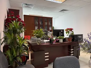 Đây là một không gian văn phòng với một chiếc bàn làm việc lớn bằng gỗ màu nâu sẫm ở trung tâm. Trên bàn có nhiều đồ vật, bao gồm một màn hình máy tính, một máy in, nhiều chồng sách và tài liệu. Bàn làm việc được trang trí bởi nhiều chậu cây cảnh: một cây thủy sinh trong bình thủy tinh, một chậu hoa xương rồng Giáng sinh màu hồng, và một chậu hoa trạng nguyên màu đỏ. Phía sau bàn là một chiếc ghế văn phòng màu đen và một tủ sách lớn bằng gỗ chứa đầy sách. Bên trái có một cây cảnh lớn được trang trí bằng hoa hồng đỏ. Bên phải có một bình hoa màu tím. Ở phía trước, có một chiếc ghế văn phòng màu xám. Tường phòng màu trắng và trần nhà có các tấm thạch cao và hệ thống đèn chiếu sáng.。（日本語訳）。これは、中央に濃い茶色の木製の大きな机があるオフィススペースです。机の上には、コンピューターのモニター、プリンター、たくさんの本や書類の山など、多くの物があります。机は、ガラスの瓶に入った水生植物、ピンク色のシャコバサボテンの鉢、そして赤いポインセチアの鉢といった、多くの観葉植物で飾られています。机の後ろには、黒いオフィスチェアと、本でいっぱいの大きな木製の本棚があります。左側には、赤いバラで飾られた大きな観葉植物があります。右側には、紫色の花瓶があります。手前には、灰色のオフィスチェアがあります。部屋の壁は白く、天井には石膏ボードと照明システムがあります。