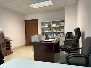 Đây là một căn phòng văn phòng. Ở giữa phòng là một chiếc bàn làm việc bằng gỗ màu nâu sẫm, trên bàn có một máy tính xách tay đang mở, một máy in, và nhiều chồng tài liệu, sổ sách. Phía sau bàn làm việc là một kệ sách lớn màu trắng, chứa đầy sách vở và hồ sơ được xếp chồng lên nhau. Có hai chiếc ghế văn phòng; một chiếc ghế giám đốc bọc da màu đen và một chiếc ghế bọc nỉ màu xám ở phía trước. Bên trái, có thể thấy một phần của một người lớn tuổi với mái tóc bạc, ngồi gần một chiếc tủ gỗ nhỏ. Căn phòng có tường sơn trắng, sàn lát gạch men màu sáng, và trần thạch cao với một đèn huỳnh quang lớn đang bật.。（日本語訳）。これはオフィスの部屋です。部屋の中央には濃い茶色の木製の事務机があり、机の上には開いているノートパソコン、プリンター、そしてたくさんの書類や本の山があります。事務机の後ろには大きな白い本棚があり、積み重ねられた本や書類でいっぱいです。オフィスチェアが2脚あります。黒い革張りの社長椅子1脚と、手前にある灰色の布張りの椅子1脚です。左側には、白髪の年配者の一部が見え、小さな木製のキャビネットの近くに座っています。部屋には白いペンキ塗りの壁、明るい色のタイル張りの床、そして大きな蛍光灯がついている石膏ボードの天井があります。