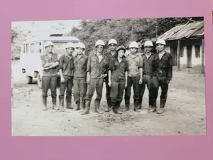 彼らは屋外の、工事現場か農村地域と思われる場所で横一列に並んでいます。ほとんどの人は、作業着と思われる、シャツ、暗い色のズボン、ブーツといった同じような服装をしています。彼らは皆、保護帽をかぶっています。女性は中央付近に立っており、微笑んでいます。彼らの後ろには、左側にミニバスのような車が、右側にはトタン屋根の簡素な家があります。一番奥には木々が鬱蒼と茂っています。この写真は、作業チームまたは労働者たちの記念撮影の雰囲気を帯びています