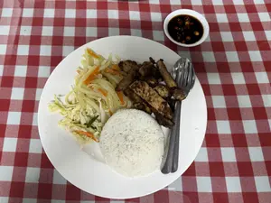 Một đĩa thức ăn được đặt trên khăn trải bàn sọc caro đỏ và trắng. Trên đĩa có một phần cơm trắng được nén thành hình tròn, thịt nướng cắt miếng, và rau xào hoặc dưa góp làm từ bắp cải và cà rốt. Bên cạnh thịt nướng có một chiếc thìa và một chiếc nĩa. Ở phía trên bên phải là một chén nước chấm nhỏ màu trắng đựng nước tương và ớt cắt lát.。（日本語訳）。赤と白のチェック柄のテーブルクロスの上に、一皿の料理が置かれています。皿の上には、丸い形に押し固められた白米、切り分けられた焼き肉、そしてキャベツと人参で作られた野菜炒めか漬物があります。焼き肉の隣には、スプーンとフォークがあります。右上には、醤油とスライスされた唐辛子が入った、小さな白いタレの小鉢があります。