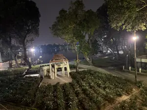 Quang cảnh một công viên ven hồ vào ban đêm. Ở phía trước là một vườn hoa hồng trắng và một công trình kiến trúc nhỏ kiểu cổ có mái ngói. Con đường đi dạo uốn lượn quanh co dưới những tán cây lớn. Phía xa là mặt hồ rộng lớn, phản chiếu ánh đèn của thành phố. Một vài người đang đi dạo ven hồ. Không gian yên tĩnh và thanh bình.。（日本語訳）。夜の湖畔公園の光景。手前には白いバラ園と、瓦屋根の小さな古い様式の建造物がある。遊歩道は大きな木々の下を蛇行している。遠くには広大な湖面が広がり、街の灯りを映している。数人が湖畔を散策している。空間は静かで穏やかだ。