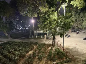 Đây là khung cảnh một công viên vào ban đêm, được chụp từ góc nhìn trên cao. Phía trước là một vườn hoa hồng với nhiều bụi cây có hoa màu trắng. Bên cạnh vườn hoa là một khoảng sân rộng, nơi có nhiều xe máy đang được đỗ. Một cây cổ thụ lớn với tán lá xum xuê được ánh đèn đường chiếu sáng rực rỡ. Ánh sáng từ các cột đèn tạo ra những vùng sáng và tối xen kẽ, mang lại một không gian yên tĩnh. Ở phía xa, có thể thấy thêm nhiều cây cối và một tòa nhà nhỏ.。（日本語訳）。これは夜の公園の光景で、上からの視点で撮られたものです。手前には、白い花をつけた茂みがたくさんあるバラ園があります。バラ園の隣には広い中庭があり、そこには多くのバイクが停められています。青々と茂った葉を持つ一本の大木が、街灯に明るく照らされています。街灯からの光が明るい部分と暗い部分を交互に作り出し、静かな空間をもたらしています。遠くには、さらに多くの木々と小さな建物が見えます。