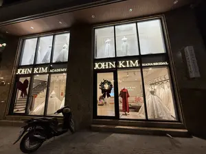 Ảnh chụp mặt tiền của một cửa hàng áo cưới vào ban đêm. Tên cửa hàng là JOHN KIM ACADEMY và JOHN KIM HANA BRIDAL được viết bằng chữ phát sáng. Bên trong các ô cửa kính lớn được chiếu sáng rực rỡ, nhiều ma-nơ-canh đang trưng bày những bộ váy cưới màu trắng với các kiểu dáng khác nhau. Ở tầng trệt, nổi bật là một chiếc váy cưới công chúa xòe rộng, đính đá lấp lánh, và một ma-nơ-canh khác mặc một bộ trang phục màu đỏ. Cửa kính được trang trí bằng một vòng hoa Giáng sinh có nơ đỏ. Phía trước cửa hàng có một chiếc xe máy đang đỗ trên vỉa hè.。（日本語訳）。夜に撮影されたウェディングドレス店の正面の写真。店名はJOHN KIM ACADEMYとJOHN KIM HANA BRIDALで、光る文字で書かれている。明るく照らされた大きなガラス窓の内側では、多くのマネキンが様々なデザインの白いウェディングドレスを展示している。1階には、きらきら光る石がちりばめられた、裾の広がったプリンセスラインのウェディングドレスが目立ち、別のマネキンは赤い衣装を着ている。ガラスのドアは赤いリボンのついたクリスマスリースで飾られている。店の前には、歩道に一台のバイクが停まっている。
