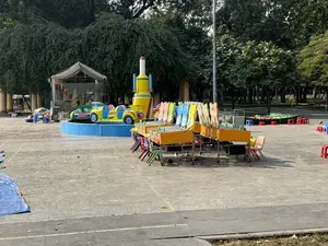 Bức ảnh chụp một khu vui chơi giải trí dành cho trẻ em được dựng lên ngoài trời, có thể là trong một công viên. Ở phía trước và trung tâm là một dãy các quầy trò chơi di động có bánh xe, được sơn màu vàng với các tấm bảng phía sau có hình động vật nhiều màu sắc. Trước mỗi quầy là những chiếc ghế nhựa nhỏ màu xanh lá, tím, hồng. Phía sau, có một vòng đu quay nhỏ với các toa xe hình ô tô màu sắc sặc sỡ như vàng, xanh dương, và một mô hình tên lửa lớn màu vàng. Bên cạnh đó là một chiếc lều trắng. Ở phía xa bên phải, có một khu vực quây lại giống như một bể bơi bơm hơi nhỏ với nhiều ghế nhựa màu đỏ và xanh dương xung quanh. Toàn bộ khu vực được bao quanh bởi cây cối xanh tươi. Sân bê tông có một ít lá rụng, và không có người nào đang chơi, tạo cảm giác vắng vẻ.。（日本語訳）。写真は、屋外、おそらく公園に設置された子供向けの遊園地を写しています。手前中央には、車輪付きの移動式ゲーム屋台が並んでおり、黄色に塗られ、後ろの看板にはカラフルな動物の絵が描かれています。各屋台の前には、緑、紫、ピンクの小さなプラスチックの椅子があります。後ろには、黄色や青などの色鮮やかな車型のゴンドラが付いた小さな観覧車と、大きな黄色のロケットの模型があります。その隣には白いテントがあります。右奥には、小さなビニールプールのような囲われたエリアがあり、その周りには赤と青のプラスチックの椅子がたくさんあります。エリア全体が青々とした木々に囲まれています。コンクリートの地面には落ち葉が少しあり、遊んでいる人はおらず、閑散とした雰囲気です。