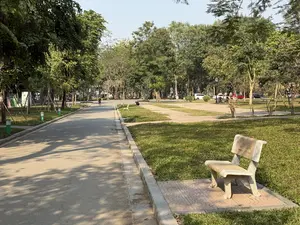 Bức ảnh chụp một khung cảnh trong công viên vào một ngày nắng đẹp. Ở phía trước, một chiếc ghế đá được đặt trên nền gạch hoa bên cạnh bãi cỏ xanh mướt. Một con đường nhỏ trải nhựa chạy dài, trên đó in bóng của những tán cây. Dọc theo con đường là những hàng cây xanh và một vài chiếc ghế đá khác. Ở phía xa, có một vài người đang đi dạo và hoạt động trong công viên. Xa hơn nữa là khu vực đỗ xe với vài chiếc ô tô. Bầu không khí trông thật yên bình và trong lành.。（日本語訳）。写真は、晴れた美しい日の公園の風景を写しています。手前には、青々とした芝生の隣、模様のあるタイルの床の上に石のベンチが置かれています。小さな舗装された道が長く伸びており、その上には木々の影が映っています。道に沿って、緑の並木といくつかの他の石のベンチがあります。遠くには、公園を散歩したり活動したりしている数人の人がいます。さらに遠くには、数台の車が停まっている駐車場があります。雰囲気はとても穏やかで清々しいです。