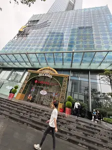 Bức ảnh chụp từ góc thấp, hướng lên một tòa nhà cao tầng hiện đại với mặt tiền bằng kính. Lối vào của trung tâm thương mại Lotte được trang trí cho dịp lễ với một cổng chào lớn màu vàng và xanh lá cây có dòng chữ 'SWEET HOLIDAYS'. Bên trong cửa kính có thể thấy các đồ trang trí như vòng quay ngựa gỗ. Ở phía trước, một thanh niên mặc áo phông trắng, quần jean đen và đeo khẩu trang đang đi bộ. Trên các bậc thang dẫn lên lối vào, có một vài người khác đang đứng và ngồi. Bầu trời có vẻ u ám.。（日本語訳）。写真はローアングルから撮影されており、ガラス張りの近代的な高層ビルを見上げています。ロッテのショッピングセンターの入り口は、祝祭日のために「SWEET HOLIDAYS」という文字が入った黄色と緑色の大きなアーチで飾られています。ガラス扉の内側には、メリーゴーランドのような装飾が見えます。手前では、白いTシャツ、黒いジーンズを履き、マスクをした一人の若者が歩いています。入り口へと続く階段の上には、他に数人が立ったり座ったりしています。空は曇っているようです。