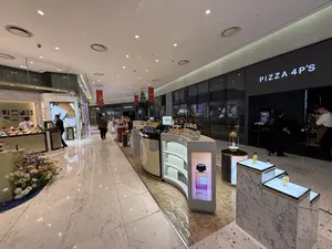 Quang cảnh bên trong một trung tâm thương mại sang trọng với sàn nhà lát đá hoa cương sáng bóng. Ở giữa lối đi là khu vực trưng bày nước hoa với nhiều quầy kệ có thiết kế hiện đại, trưng bày các sản phẩm nước hoa. Phía sau là nhà hàng PIZZA 4P'S với biển hiệu phát sáng. Có một vài người đang đi lại và mua sắm. Trần nhà được trang trí bằng các dải băng màu đỏ cho dịp lễ hội.。（日本語訳）。光沢のある大理石の床を持つ、豪華なショッピングセンターの内部の光景。通路の中央には、モダンなデザインの多くの棚やカウンターがある香水の展示エリアがあり、香水製品が陳列されている。後方には、光る看板のあるレストランPIZZA 4P'Sがある。数人が行き来し、買い物をしている。天井は祝祭行事のために赤いリボンで飾られている。