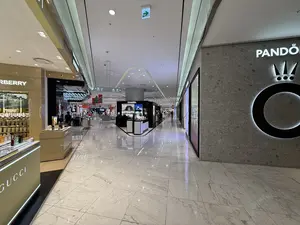 Đây là hình ảnh một hành lang rộng rãi và sang trọng bên trong một trung tâm thương mại hoặc khu mua sắm miễn thuế. Sàn nhà được lát gạch men trắng sáng bóng, phản chiếu ánh đèn từ trần nhà cao. Dọc hai bên hành lang là các cửa hàng và quầy trưng bày của những thương hiệu nổi tiếng. Bên trái, có thể thấy rõ quầy hàng của GUCCI và BURBERRY với nhiều sản phẩm nước hoa được trưng bày. Ở giữa lối đi là một quầy hàng độc lập của CHANEL. Phía bên phải là bức tường có logo lớn của thương hiệu trang sức PANDORA. Không gian rất sáng sủa, hiện đại và có một vài người đang mua sắm ở phía xa.。（日本語訳）。これは、ショッピングセンターまたは免税店の中にある、広々として豪華な廊下の画像です。床は光沢のある白いタイルで敷き詰められており、高い天井からの光を反射しています。廊下の両脇には、有名ブランドの店舗や陳列カウンターが並んでいます。左側には、GUCCIとBURBERRYのカウンターがはっきりと見え、多くの香水製品が陳列されています。通路の中央には、CHANELの独立したカウンターがあります。右側には、ジュエリーブランドPANDORAの大きなロゴがある壁があります。空間はとても明るくモダンで、遠くで数人が買い物をしています。