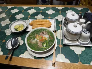 Một bữa ăn phở Việt Nam được bày biện thịnh soạn trên một chiếc bàn có mặt gạch hoa màu xanh lá cây với họa tiết hình hoa bốn cánh. Trung tâm của bức ảnh là một tô phở bò bốc khói, với nước dùng trong, thịt bò tái, bánh phở trắng và rất nhiều hành lá, hẹ thái nhỏ, cùng vài lát ớt. Bên cạnh là một đĩa quẩy vàng óng, một bát nhỏ đựng chanh cắt miếng để vắt thêm vào phở. Ngoài ra còn có một bộ ấm và hũ đựng gia vị bằng sứ trắng, được trang trí bằng hoa văn màu xanh đen, đặt gọn gàng trên một chiếc khay. Một đôi đũa tre và một chiếc thìa inox cũng được chuẩn bị sẵn sàng cho bữa ăn.。（日本語訳）。四枚の花びらの模様がある緑色の花柄タイルが張られたテーブルの上に、ベトナムのフォーの食事が豪華に盛り付けられています。写真の中心には湯気の立つ牛肉のフォーの丼があり、透明なスープ、レアな牛肉、白いフォーの麺、そしてたくさんの刻んだネギとニラ、それに数枚の唐辛子のスライスが入っています。隣には黄金色の揚げパン（クァイ）の皿と、フォーに絞るためのカットライムが入った小さなボウルがあります。その他にも、濃い青色の模様で装飾された白い磁器の急須と調味料入れのセットが盆の上にきちんと置かれています。竹の箸一膳とステンレスのスプーンも食事のために用意されています。