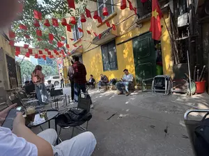 PHỞ THÌN。150。21。Khung cảnh một quán cà phê vỉa hè trong một con ngõ nhỏ ở Hà Nội. Tòa nhà có bức tường màu vàng cũ kỹ, cửa sổ lá sách màu xanh. Phía trên, nhiều dây cờ đỏ sao vàng và cờ Đảng được giăng ngang, tạo không khí lễ hội. Một lá cờ Tổ quốc lớn được treo bên cạnh cửa ra vào. Nhiều người đang ngồi trên những chiếc ghế xếp đơn giản, thưởng thức đồ uống và thư giãn. Một số người đang chăm chú vào điện thoại di động. Trên bàn ở phía trước có một ly cà phê nâu đá và một ly trà. Không gian mang đậm nét đời thường, gần gũi và đặc trưng của cuộc sống đường phố Việt Nam.。（日本語訳）。フォー・ティン。150。21。ハノイの小さな路地にある歩道のカフェの光景。建物は古びた黄色の壁で、緑色のルーバー窓がある。上には、金星紅旗と党旗の多くの旗が横に張られ、お祭りの雰囲気を醸し出している。多くの人々が簡素な折りたたみ椅子に座り、飲み物を楽しんだりリラックスしたりしている。何人かの人々は携帯電話に夢中になっている。手前のテーブルには、アイスミルクコーヒーが一杯とお茶が一杯ある。その空間は、ベトナムのストリートライフの日常的で、親しみやすく、特徴的な雰囲気に満ちている。