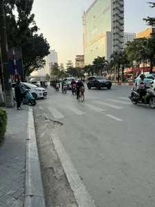 Một con đường đông đúc ở thành phố vào buổi chiều tà. Dòng người và phương tiện gồm nhiều xe máy, ô tô, và một người đi xe đạp đang lưu thông trên đường có vạch kẻ cho người đi bộ. Bên vỉa hè, hai người đàn ông đang đứng nói chuyện cạnh một chiếc xe máy. Phía sau là những hàng cây xanh và các tòa nhà cao tầng hiện đại, trong đó có một tòa nhà văn phòng lớn với mặt tiền bằng kính. Ánh nắng vàng của hoàng hôn chiếu lên các tòa nhà và con đường.。（日本語訳）。夕暮れ時の街の混雑した道。多くのバイク、車、そして自転車に乗った一人からなる人々と乗り物の流れが、横断歩道のある道路を通行している。歩道脇では、二人の男性がバイクの隣で立ち話をしている。後方には緑の並木と近代的な高層ビルがあり、その中には正面がガラス張りの大きなオフィスビルがある。夕日の黄金色の光が、ビル群と道路を照らしている。