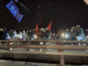 Một đám đông người đi xe máy đang dừng lại trên đường vào ban đêm. Nhiều người đội mũ bảo hiểm và một số người cầm cờ đỏ sao vàng của Việt Nam. Khung cảnh có vẻ là một buổi diễu hành hoặc ăn mừng, có thể là sau một trận bóng đá. Ánh sáng từ đèn pha xe máy chiếu sáng con đường. Phía trước là một lan can kim loại cũ.。（日本語訳）。夜、バイクに乗った人々の集団が路上で停止しています。多くの人がヘルメットをかぶっており、何人かはベトナムの金星紅旗を手に持っています。この光景はパレードかお祝いのようで、おそらくサッカーの試合の後でしょう。バイクのヘッドライトの光が道路を照らしています。手前には古い金属製の手すりがあります。