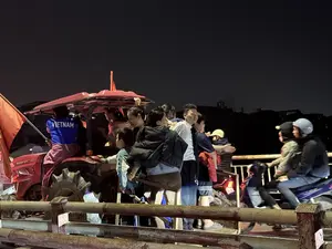 VIETNAM. Khung cảnh ban đêm, một nhóm người gồm cả người lớn và trẻ em đang tụ tập trên một chiếc máy cày màu đỏ, có vẻ như đang ăn mừng. Một người ngồi trên xe mặc áo màu xanh lam, sau lưng có in chữ 'VIETNAM' và đang cầm một lá cờ đỏ. Những người khác đứng trên xe và xung quanh, tạo nên một không khí sôi động.。（日本語訳）。ベトナム。夜の光景、大人と子供を含む一団が赤いトラクターの上に集まっており、祝っているようです。車の上に座っている一人が青いシャツを着ており、背中には「VIETNAM」の文字がプリントされ、赤い旗を持っています。他の人々は車の上や周りに立っており、活気のある雰囲気を醸し出しています。