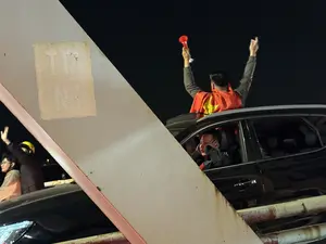 T17 NI. Cảnh các cổ động viên ăn mừng trên đường phố vào ban đêm. Một người đàn ông khoác cờ Việt Nam đứng trên nóc xe ô tô, giơ cao hai tay, một tay cầm chiếc kèn màu đỏ. Một người khác ngồi trong xe thò đầu ra ngoài cửa sổ, hai tay ôm đầu. Ở phía trước, trên một trụ cột có dán ký hiệu 'T17 NI'.。（日本語訳）。T17 NI。夜の路上で祝うサポーターたちの光景。ベトナム国旗を羽織った男性が車の屋根の上に立ち、両手を高く上げ、片手には赤いラッパを持っている。別の人物が車の中に座り、窓から頭を出し、両手で頭を抱えている。前方には、柱に「T17 NI」という記号が貼られている。