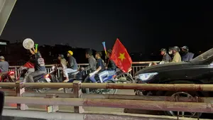 Một nhóm người hâm mộ đang đi xe máy trên đường phố vào ban đêm, có vẻ như đang ăn mừng chiến thắng bóng đá. Một người cầm lá cờ đỏ sao vàng của Việt Nam, trong khi những người khác cầm kèn và một người giơ cao một chiếc vung nồi. Mọi người đều đội mũ bảo hiểm, tạo nên một khung cảnh náo nhiệt và đầy phấn khích trên một cây cầu.。（日本語訳）。ファンのグループが夜の路上をバイクで走っており、サッカーの勝利を祝っているようです。一人はベトナムの金星紅旗を掲げ、他の人たちはクラクションを持ち、一人は鍋の蓋を高く掲げています。全員がヘルメットを着用しており、橋の上で賑やかで興奮に満ちた光景を作り出しています。