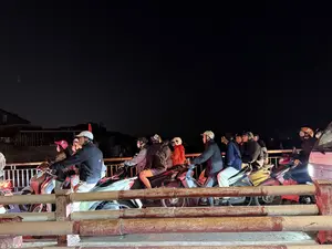 Cảnh tượng giao thông đông đúc vào ban đêm. Một hàng dài người đi xe máy đang dừng lại, có thể là do tắc đường trên một cây cầu. Hầu hết mọi người đều đội mũ bảo hiểm và mặc áo khoác. Bầu trời tối đen, phía xa là bóng của các tòa nhà. Ở phía trước, có một lan can bằng kim loại cũ được sơn màu đỏ và trắng.。（日本語訳）。夜の混雑した交通風景。バイクに乗った人々の長い列が停止している。おそらく橋の上での交通渋滞のためだろう。ほとんどの人がヘルメットをかぶり、ジャケットを着ている。手前には、赤と白に塗られた古い金属製の手すりがある。