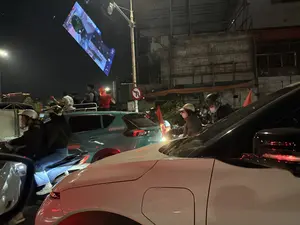 Cảnh tượng trên đường phố vào ban đêm với dòng xe cộ đông đúc gồm ô tô và xe máy. Nhiều người đang tham gia giao thông, một số đội mũ bảo hiểm và đeo khẩu trang. Có một nhóm người đứng trên một chiếc xe, trong đó có người mặc áo trắng số 18. Một số người cầm cờ đỏ, tạo không khí như một buổi ăn mừng. Phía trên có một màn hình LED lớn đang chiếu hình ảnh và một biển báo giao thông cấm quay đầu xe.。（日本語訳）。夜の路上、自動車やバイクで混雑する交通の光景。多くの人々が通行しており、一部はヘルメットをかぶり、マスクを着用している。一台の車の上に立っている人々のグループがおり、その中には18番の白いシャツを着た人がいる。一部の人々は赤い旗を持っており、お祝いのような雰囲気を醸し出している。上方には映像を映している大きなLEDスクリーンと、Uターン禁止の交通標識がある。