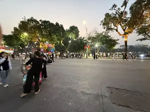 Bức ảnh chụp cảnh một con phố đi bộ đông đúc vào buổi chập choạng tối. Không khí rất nhộn nhịp với nhiều người qua lại. Ở phía bên trái, một người bán hàng rong đang đẩy một chiếc xe chở đầy bóng bay nhiều màu sắc và hình thù, bao gồm cả hình hươu cao cổ. Mọi người, bao gồm cả người lớn và trẻ em, đang đi dạo trên đường. Hai bên đường là những hàng cây xanh lớn, và đèn đường đã bật sáng, chiếu rọi không gian. Con đường nhựa rộng rãi, và ở phía xa có thể thấy bóng của các tòa nhà và cây cối.。（日本語訳）。写真は、夕暮れ時の混雑した歩行者天国の光景を写しています。多くの人が行き交い、雰囲気はとても賑やかです。左側では、露天商がキリンの形を含む、様々な色や形の風船でいっぱいのカートを押しています。大人も子供も含む人々が、通りを散歩しています。道の両側には大きな緑の並木があり、街灯はすでに点灯し、空間を照らしています。アスファルトの道は広々としており、遠くには建物や木々の影が見えます。